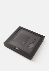 Set di portafoglio in pelle nera e portachiavi in una scatola nera. Il portafoglio presenta un logo impresso; il portachiavi ha un design con lettere intrecciate.