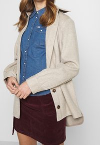 Beige ullblandning kofta med bruna knappar, lager på lager över en blå denimskjorta, kombinerad med en vinröd kordkjol.