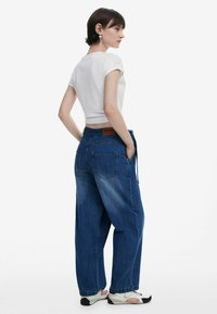 Blå denimbyxor med vida ben och ljus tvätt, med bakfickor och en brun logotypchans på midjan. Kombineras med en vit topp.
