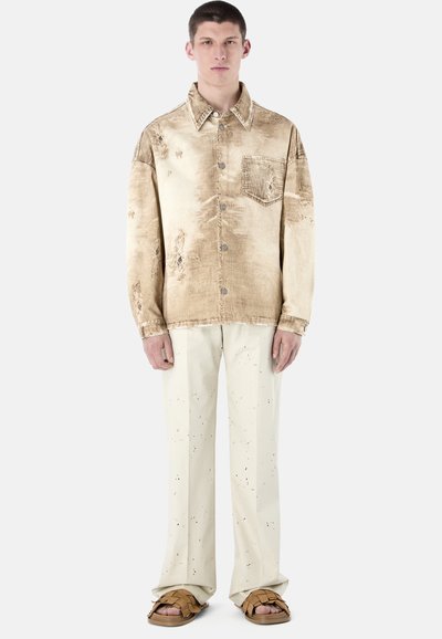 Un jeune homme debout portant une chemise beige usée à boutons, un pantalon crème moucheté et des sandales tressées marron sur un fond blanc.