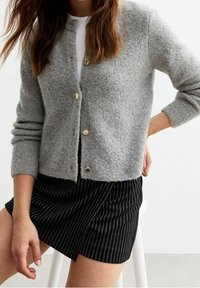 Grauer Wollcardigan mit goldenen Knöpfen, langen Ärmeln, strukturiertem Stoff, getragen über einem weißen Oberteil und einem gestreiften schwarzen Rock. Lässiges, geschichtetes Outfit.