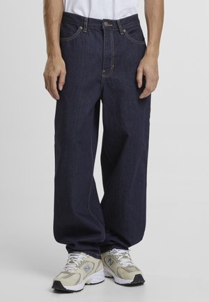 Personne portant un jean en denim bleu foncé ample et des baskets New Balance beiges avec des lacets blancs, debout devant un fond gris uni.