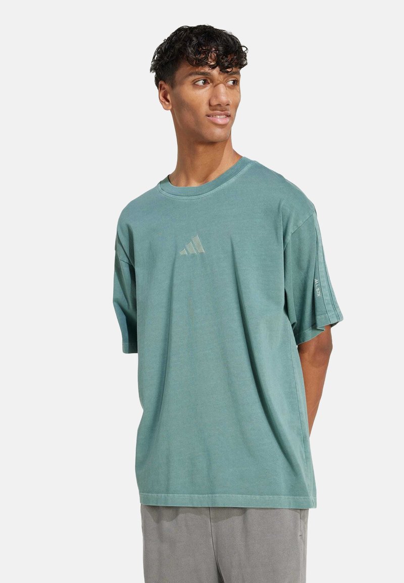 Tyrkysové bavlněné tričko s krátkým rukávem, kulatým výstřihem a světle zeleným logem Adidas na hrudi. Hladká textura a volný střih.