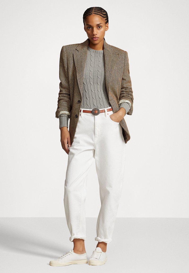 Brauner Fischgrät-Blazer, grauer Zopfpullover, weiße hochgekrempelte Hose und helle Sneakers. Ledergürtel mit einer runden Metallschnalle.