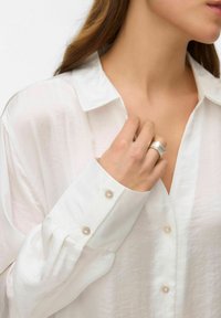 White, long-sleeve shirt with a satin finish, featuring soft buttons. A silver ring is worn on the hand, enhancing the minimalist aesthetic.