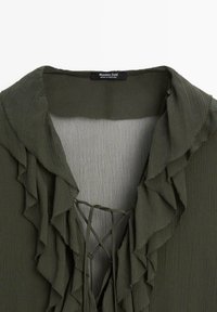 Blouse vert foncé avec un col froncé et un devant froncé, en tissu léger et texturé, avec un détail de cordon au niveau du décolleté. Étiquette de marque visible.