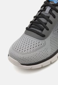 Scarpa atletica grigia con tomaia in rete e accenti neri. Presenta un collare imbottito, lacci neri e una suola bianca flessibile. Superficie testurizzata.