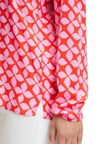 Chemise à manches longues rouge vif avec un motif de feuilles roses, portée avec un pantalon blanc, montrant un poignet et une main détendue sur le côté.