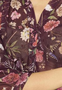 Blusa trasparente bordeaux con motivo floreale, accentuata da bottoni rosa, dotata di colletto e maniche arrotolate con una finitura texturizzata.