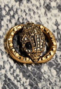 Fibbia decorativa in tono dorato con design di teschio adornato da strass neri e dorati, su uno sfondo strutturato grigio e a crema con motivo.
