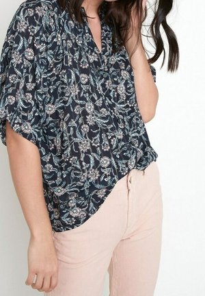 Woman wearing a black floral blouse with short sleeves and light pink high-waisted pants, posing against a white background.