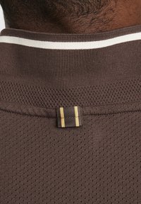 Hnedý látkový golier s textúrovaným sieťovaným dizajnom, obsahujúci biely pruh a prišitú nášivku s dvoma zlatými pruhmi pre detail.