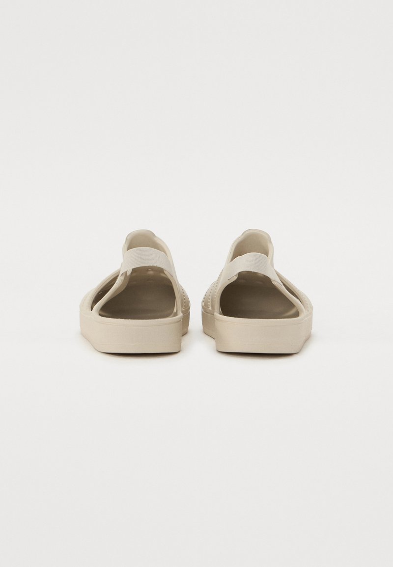 Beige Sandalen mit offenem Rücken, einem horizontalen Riemen über dem Mittelfuß, strukturiertem Sohlen und kleinen runden Perforationen an den Seiten.