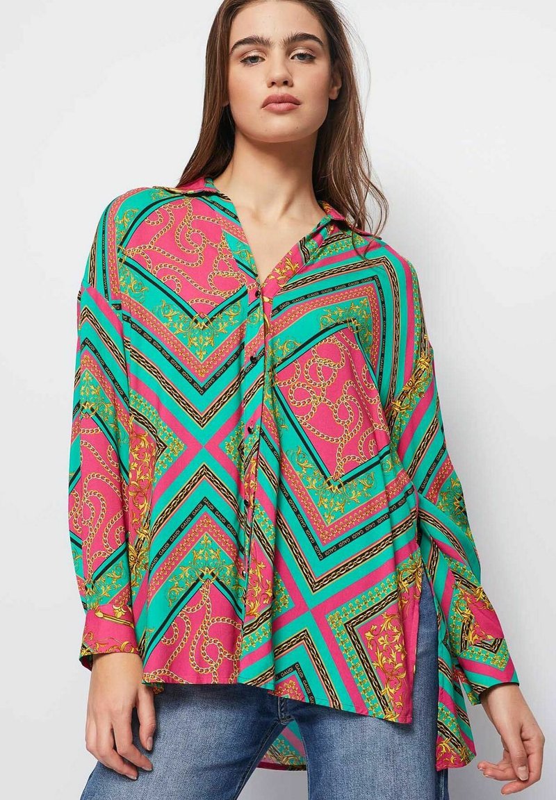 Blusa abotonada colorida con un patrón de zigzag en rosa y turquesa, acentos de cadena dorada, y mangas largas con aberturas laterales.