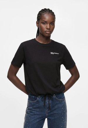 Γυναίκα με πλεξούδες, που φοράει μαύρο t-shirt Karl Lagerfeld Jeans και μπλε τζιν με μικρά ασημένια τρουκ στις τσέπες και τα πόδια.