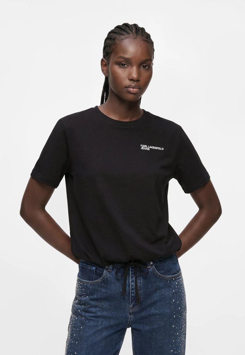 Γυναίκα με πλεξούδες, που φοράει μαύρο t-shirt Karl Lagerfeld Jeans και μπλε τζιν με μικρά ασημένια τρουκ στις τσέπες και τα πόδια.