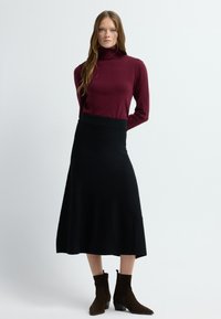 Bordeaux coltrui gecombineerd met een zwarte, knie-lange rok. Draagt bruin suède enkellaarsjes met een lage hak, tegen een lichte achtergrond.