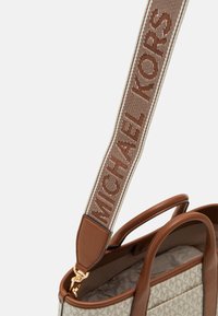 Kožená kabelka v tehlovej farbe s textúrovaným pláteným telom. Obsahuje tkaný popruh s nápisom "MICHAEL KORS" v hnedej, zlatú kovania a otvorený dizajn vrchu.