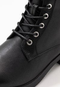 Černé kotníkové boty vyrobené z texturované kůže, s kovovými očky, plochými šňůrkami a robustní podrážkou, s viditelnými švy.