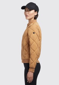 Gequilte tan bomberjack met ribbelmanchetten. Beschikt over een ronde kraag en een zwarte accentknop op de mouw. Gecombineerd met zwarte legging.