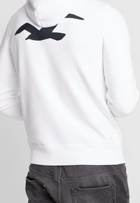 Vit hoodie med en stor svart fågelsilhuett på baksidan, tillverkad av mjukt tyg med avslappnad passform och ribbade ärmslut.