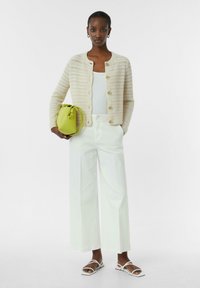 Beige gebreide cardigan met grote knopen, witte tanktop, brede witte broek en groene emmer tas. Sandalen met witte banden.
