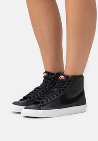 Czarne sneakersy z wysoką cholewką wykonane z gładkiej skóry, z teksturowanymi zamszowymi akcentami, białą gumową podeszwą oraz wyraźnym logo Nike na języku.