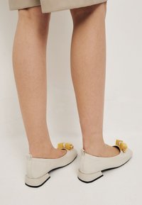 Ballerines en cuir beige avec un petit talon carré, dotées d'un détail en nœud jaune sur le bout. Surface lisse avec des accents de couture minimes.