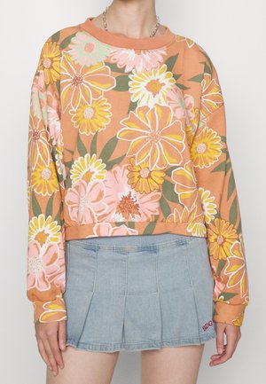 Person iført orange sweatshirt med stort lyserødt, gult og hvidt blomsterprint og lyseblå plisseret denim mininederdel.