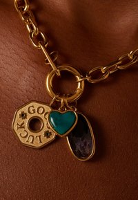 Gouden ketting met drie bedels: een gouden achthoekige hanger met "GOOD LUCK", een turquoise hart en een paarse steen.