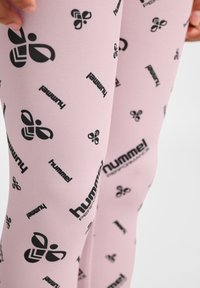Różowe legginsy wykonane z elastycznego materiału, z powtarzającym się czarnym wzorem logo. Projekt zawiera abstrakcyjne kształty i tekst.