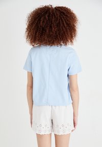 Person med krøllet hår iført en lys blå kortærmet skjorte og hvide øjenlåg lace shorts, stående med ryggen til mod en ensfarvet baggrund.