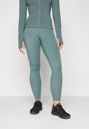 Långärmad figurnära topp och leggings i dämpad teal, tillverkade av elastiskt material. Inkluderar tumhål och kombineras med svarta sportskor.