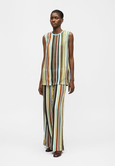 Γυναίκα που στέκεται φορώντας αμάνικη μπλούζα και παντελόνι με φαρδιά μπατζάκια, με κάθετες ρίγες σε αποχρώσεις μπλε, κίτρινου, καφέ, μαύρου και λευκού.