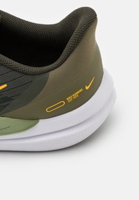 Basket de course Nike avec une tige en maille vert olive, des accents gris, une semelle blanche et des détails de marque orange, présentant un design épuré et des surfaces texturées.