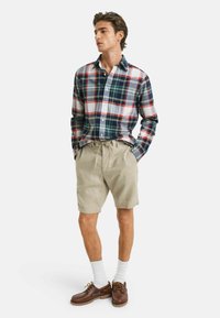 Karierte Hemd in Marineblau, Rot und Weiß; khakifarbene Shorts; braune Schuhe; weiße Socken. Merkmale sind Knöpfe, lange Ärmel und lockere Passform.