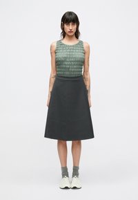 Haut sans manches en tissu vert texturé, associé à une jupe trapèze gris foncé. Le mannequin porte des chaussettes grises et des baskets blanches.