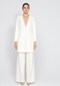 Biały skrojony garnitur z długim żakietem i szerokimi spodniami. Gładki materiał, zapięcie na jeden guzik i klapy notcha.
