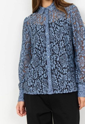 Femme portant un chemisier boutonné en dentelle florale bleue transparente à manches longues avec col, associé à un pantalon noir, vue du cou aux hanches.