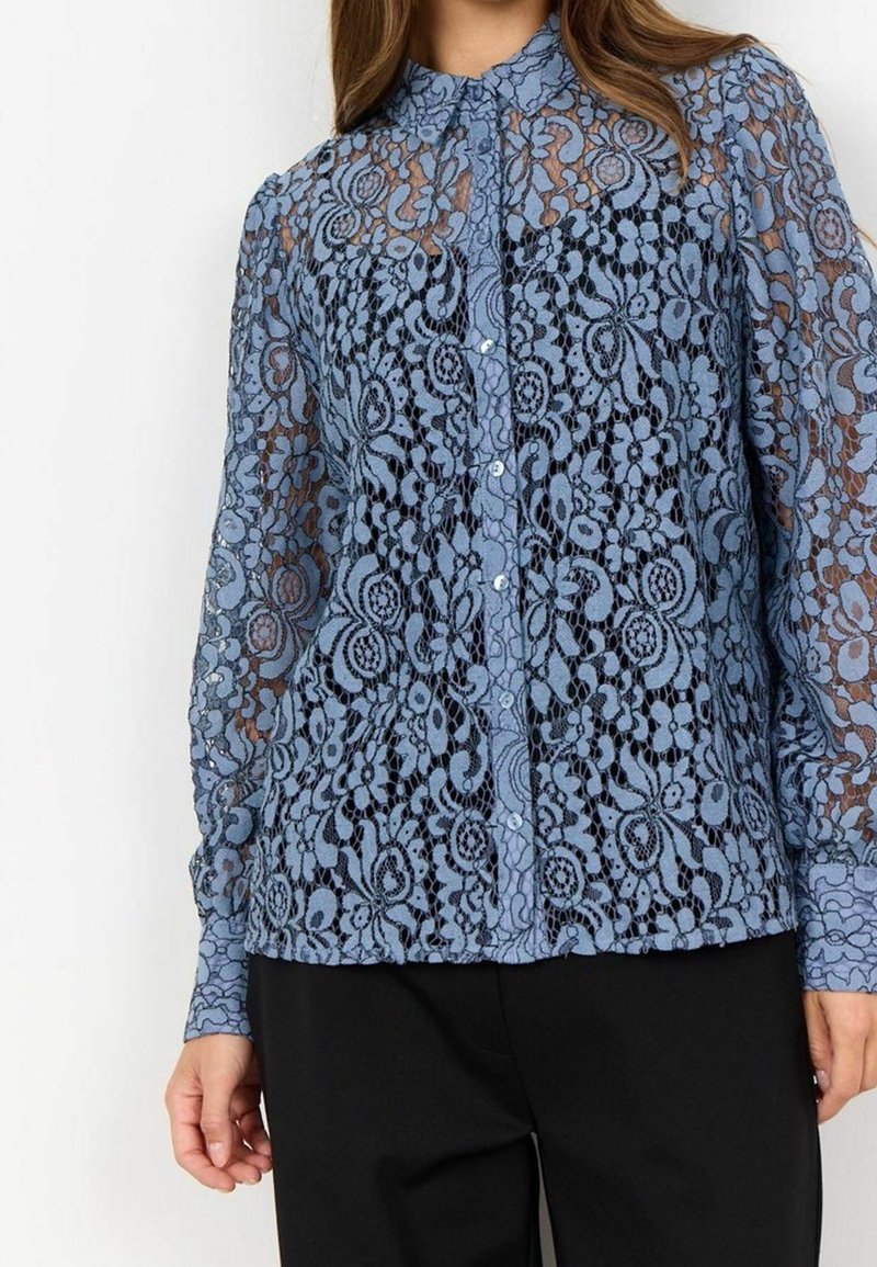 Femme portant un chemisier boutonné en dentelle florale bleue transparente à manches longues avec col, associé à un pantalon noir, vue du cou aux hanches.