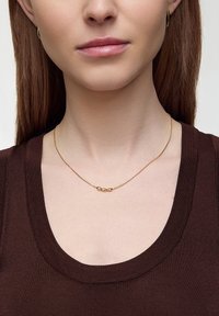 Złoty naszyjnik z trzema zaokrąglonymi akcentami na cienkim łańcuszku, noszony z ciemnobrązowym dekoltem, podkreślający jego delikatny design i metaliczny blask.