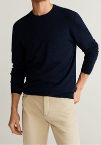 Muž v tmavě modrém svetru s dlouhým rukávem a béžových kalhotách, stojící s jednou rukou v kapse na jednoduchém světlém pozadí.