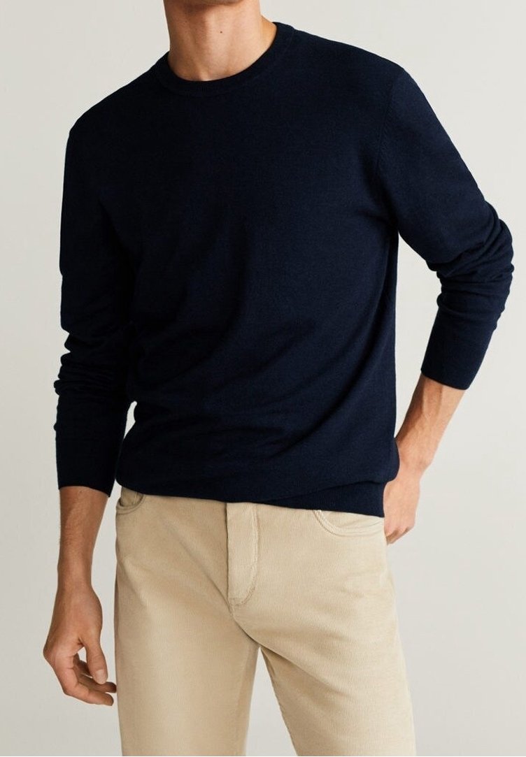 Muž v tmavě modrém svetru s dlouhým rukávem a béžových kalhotách, stojící s jednou rukou v kapse na jednoduchém světlém pozadí.