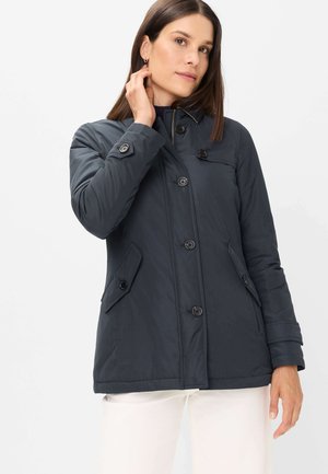 Frau trägt einen dunkelblauen, geknöpften Mantel mit Klappentaschen und weiße Hose, berührt ihren Hals vor einem schlichten weißen Hintergrund.