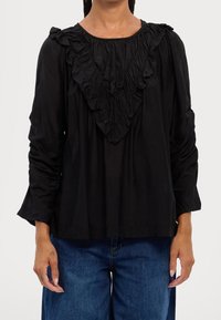 Blusa nera con scollatura arricciata, maniche lunghe e spalle arricciate. Abbinata a jeans in denim blu, che mostrano una vestibilità comoda.