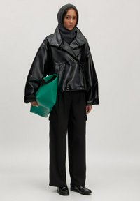 Zwarte leren double-breasted jas, brede zwarte broek en een groene tas. Model draagt een donkere gebreide capuchon. Ensemble heeft een gestructureerd ontwerp.