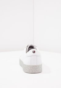 Sneaker in pelle bianca con suola argento scintillante, caratterizzata da un accento con logo rotondo sul retro. Texture liscia e design minimalista.