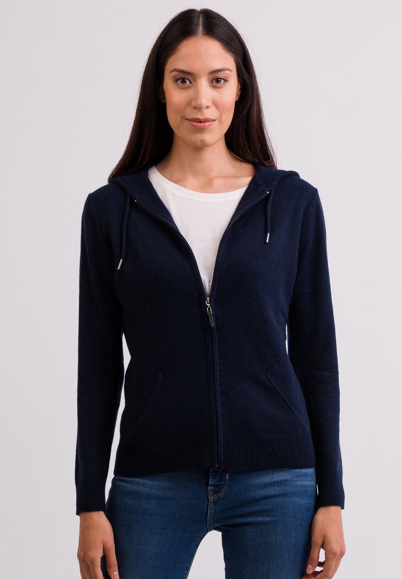 Mörkblå zip-up hoodie tillverkad av ett mjukt stickat tyg med en framdragbar dragkedja, sidofickor och snörningsdetaljer vid huvan.