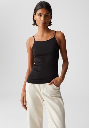 Woman with short dark hair wearing a black spaghetti strap top and off-white pants, standing with one hand in pocket against gray background.