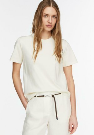 Młoda kobieta z długimi falującymi włosami, ubrana w kremowy top z krótkim rękawem i pasujące spodnie z cienkim ciemnym plecionym paskiem.
